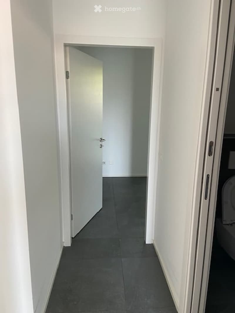 A narrow, clean hallway with white walls and dark tiled flooring, featuring an open door leading to another room.