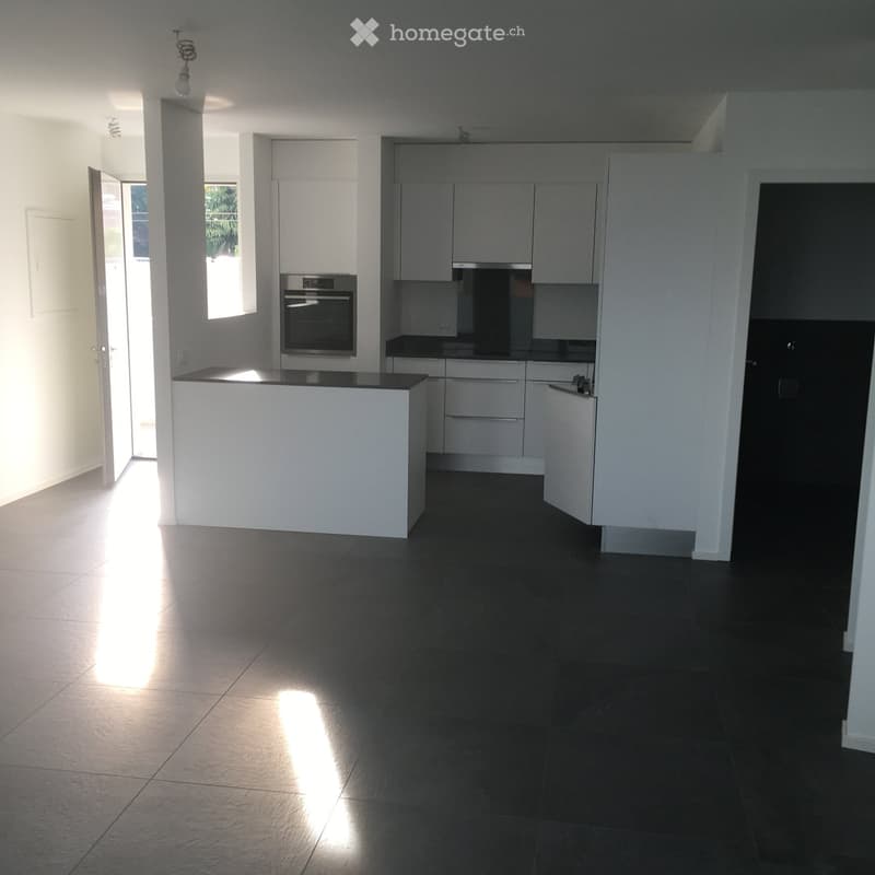 Modern kitchen with white cabinets, black countertops, and a central island. The floor is dark tiled, and natural light streams in from a window on the left.