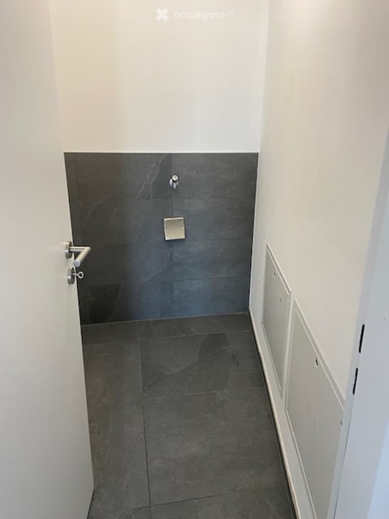A narrow hallway with white walls, dark grey tiled floor, and a small shower stall visible at the end.
