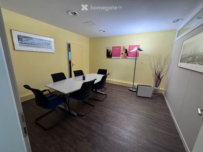 Meeting table with six chairs, white walls, yellow walls, pictures on the wall, a floor lamp, a potted plant, wooden flooring