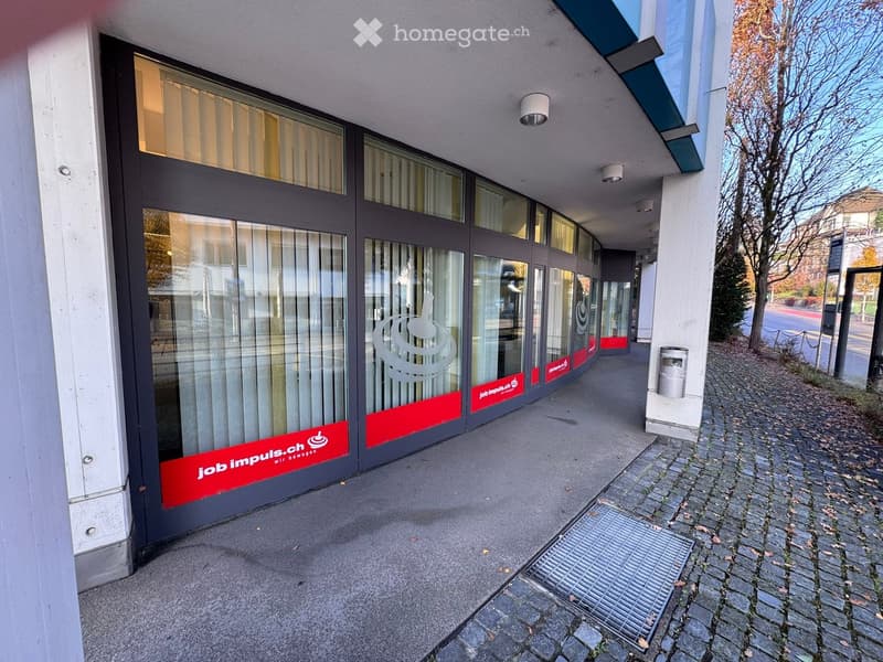 Modern building exterior with glass doors and windows, red and white branding, security cameras on the ceiling, and brick sidewalk.