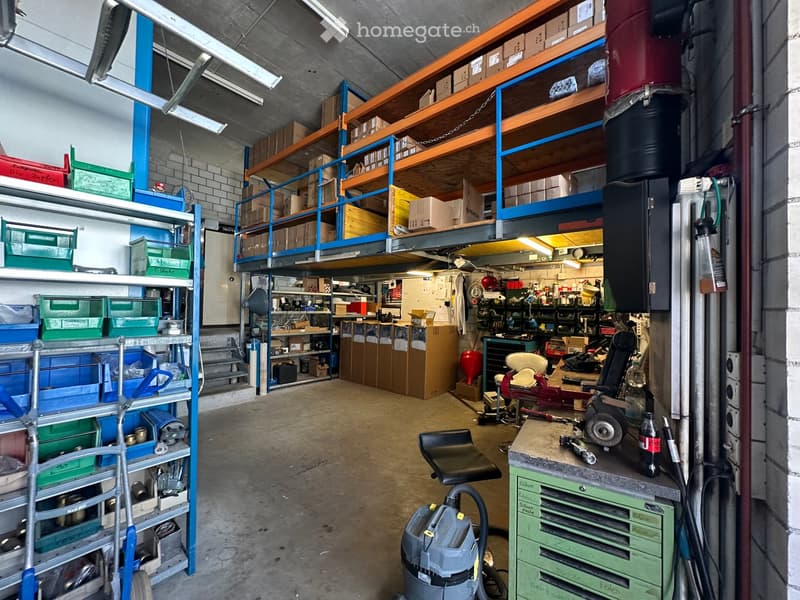 A spacious workshop with metal shelving units, tools, a workbench, a chair, and a vacuum cleaner. The area includes a lofted storage area with more boxes and tools.