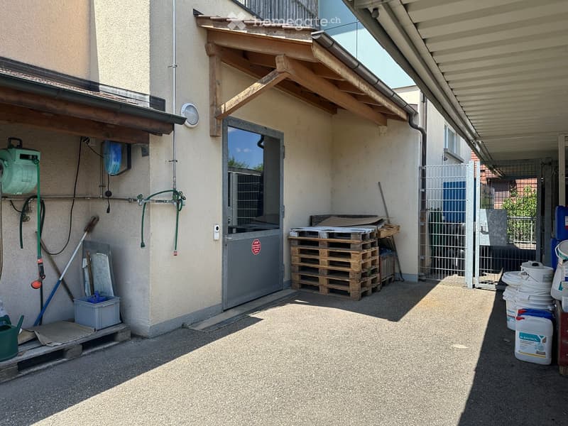 Outdoor area with a covered entrance, stacked pallets, storage containers, and a hose reel mounted on the wall.