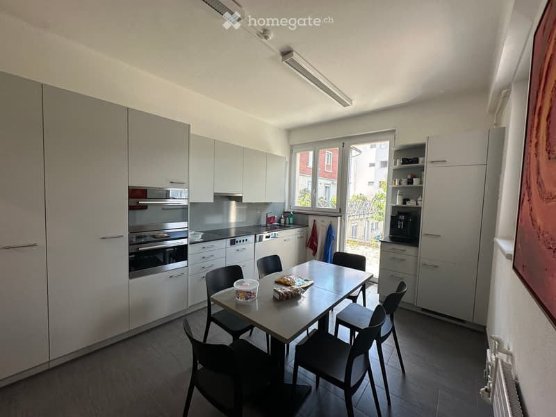 Modern kitchen with grey cabinets, stainless steel appliances, a dining table with chairs, and a window overlooking a balcony.