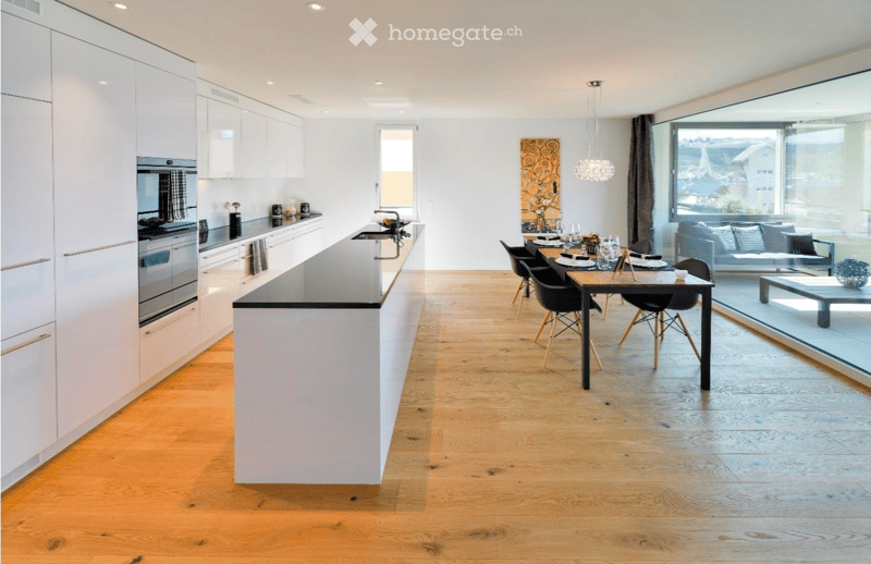 Modern kitchen and dining area with white cabinets, a central island, and a dining table with chairs. The space features wooden flooring, large windows, and a view of the outdoor area.