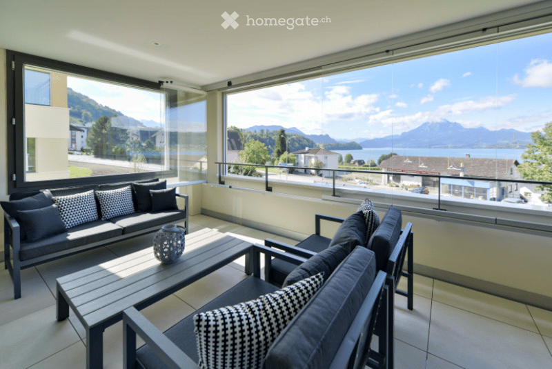 Modern balcony with large windows offering scenic mountain and lake views, furnished with a sofa, chairs, and a coffee table.