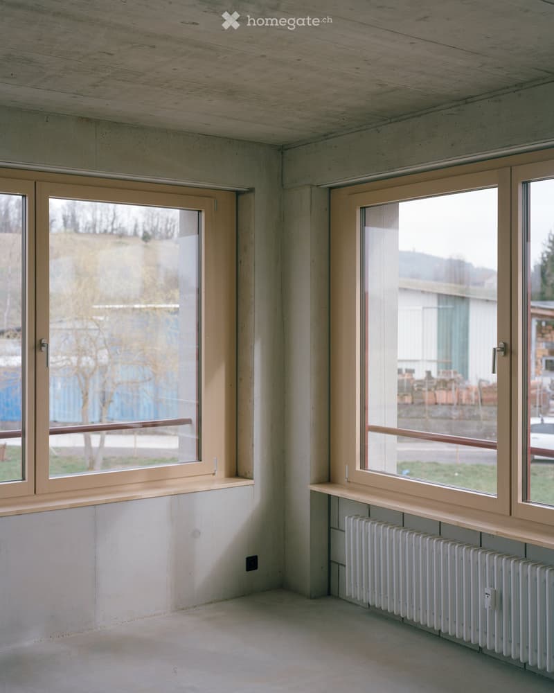 Empty room with concrete walls and floor, large windows with wooden frames, radiator, and visible electrical outlet.