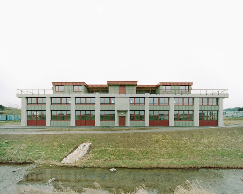 Modern two-story building with green siding, red doors, and large windows, situated near a river with a grassy bank.