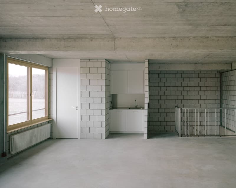 Minimalist unfurnished room with white tiled walls, concrete ceiling, large window, white cabinetry, and a metal staircase railing.