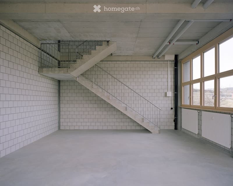 Empty room with white tiled walls, concrete floor, staircase leading to an upper level, large windows, and radiators along the wall.