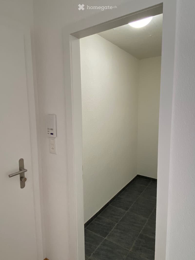 A narrow hallway with white walls, dark tiled floor, and a ceiling light fixture visible at the end.
