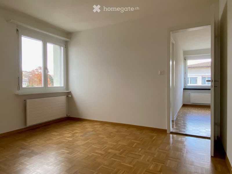 Empty room with wooden flooring, white walls, and a window with a radiator below it. An open doorway leads to another room with similar flooring and a window.