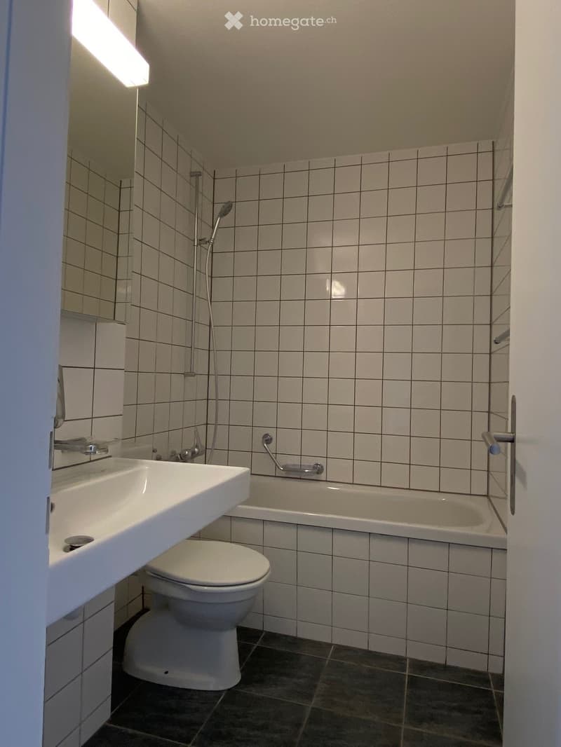 Bathroom with white tiled walls, a sink, toilet, bathtub, and a showerhead. The space is compact with dark tiled flooring.