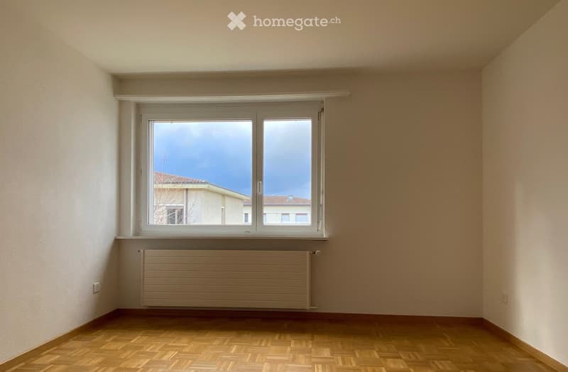 Empty room with wooden flooring, white walls, a large window with a view of a neighboring building, and a radiator below the window.