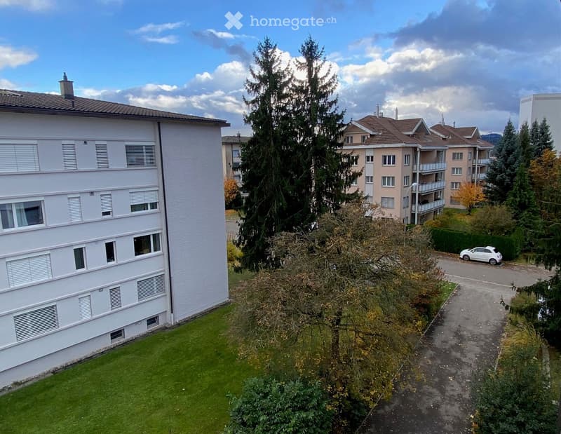 A multi-story residential building with white walls and multiple windows, surrounded by greenery and other buildings in the background.