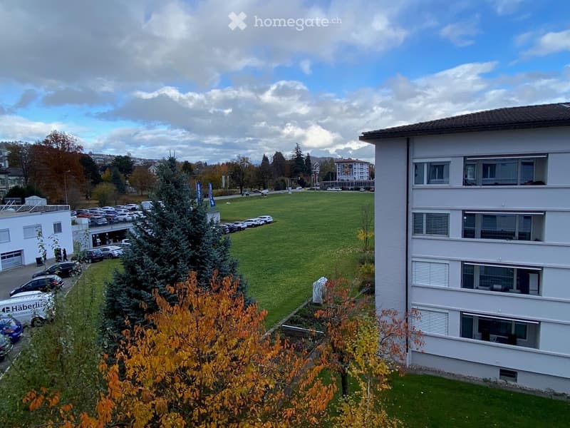 Multi-story white building with multiple windows, surrounded by a grassy area, parking lot, and trees.