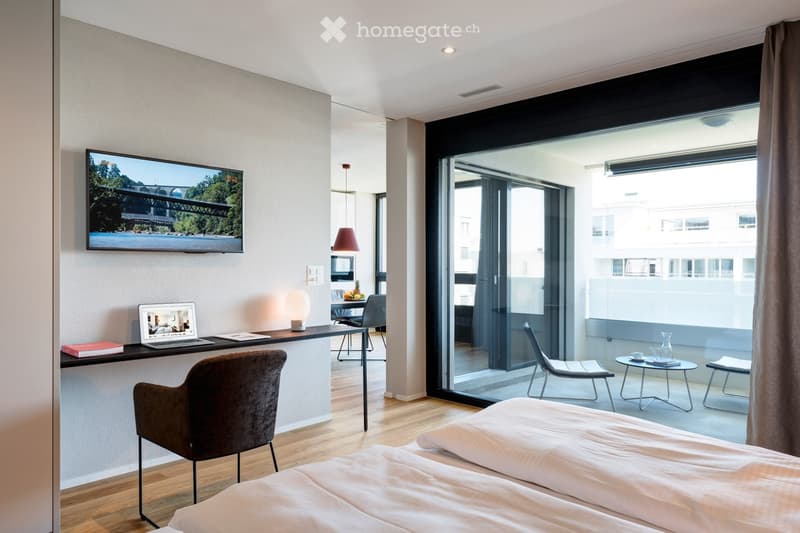 Modern bedroom with a double bed, a desk with a laptop, a chair, a lamp, a TV screen, and a small balcony area with a table and chairs.