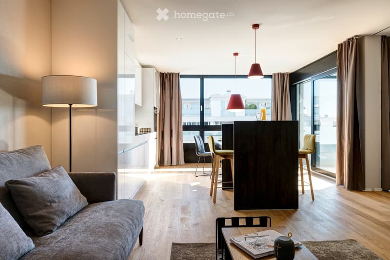 Modern living room with a grey sofa, coffee table, bar counter, and floor lamp. Large windows with curtains and a view of the city.