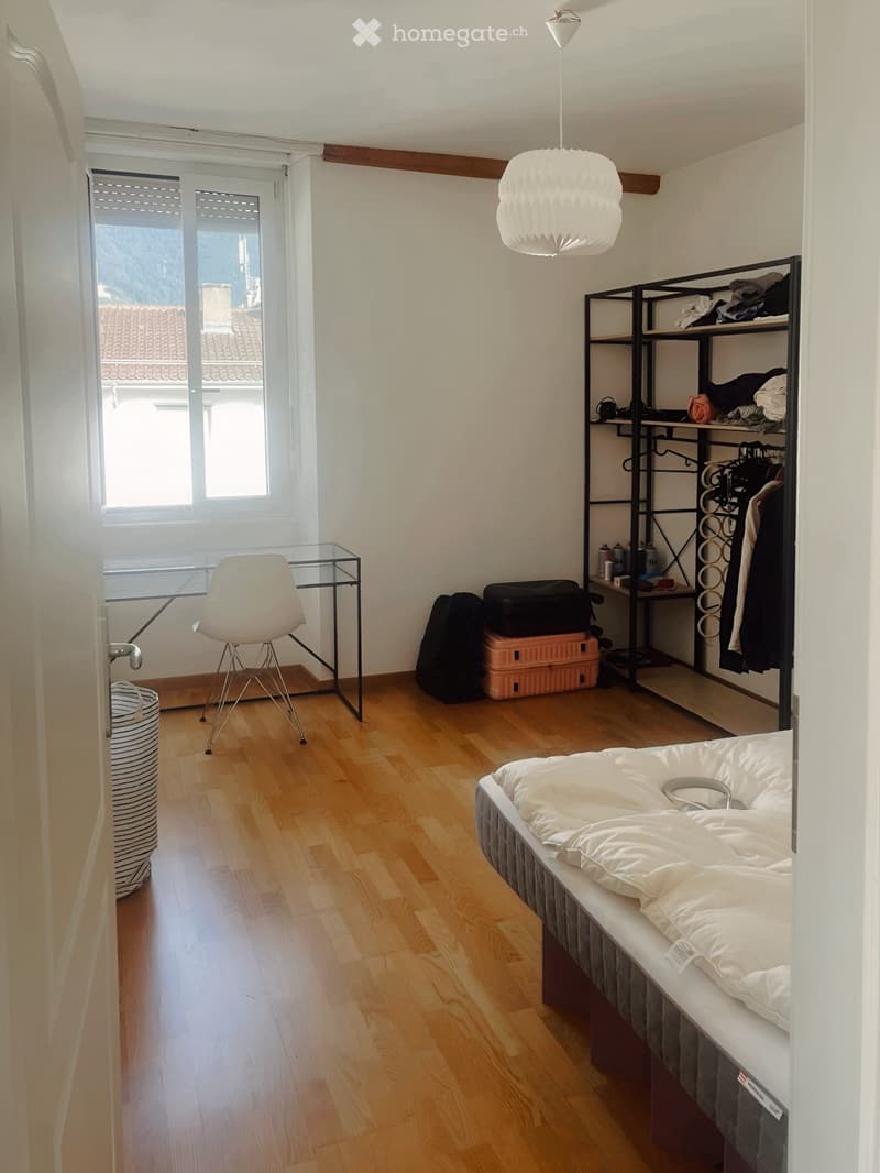 A small, well-lit bedroom with wooden flooring, a bed with white bedding, a modern chair, a shelf with folded clothes, and a small table with a lamp.