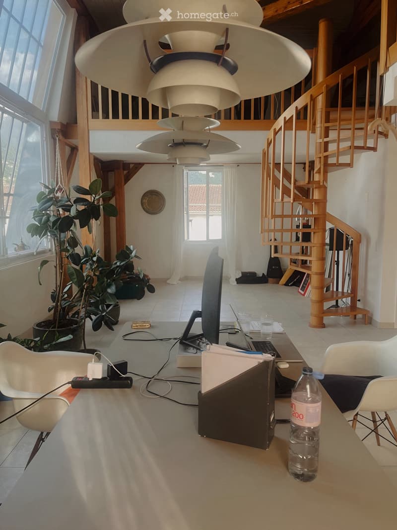 A modern, open-plan living and working space with a large staircase, white chairs, a desk with a laptop, and a water bottle on the table.
