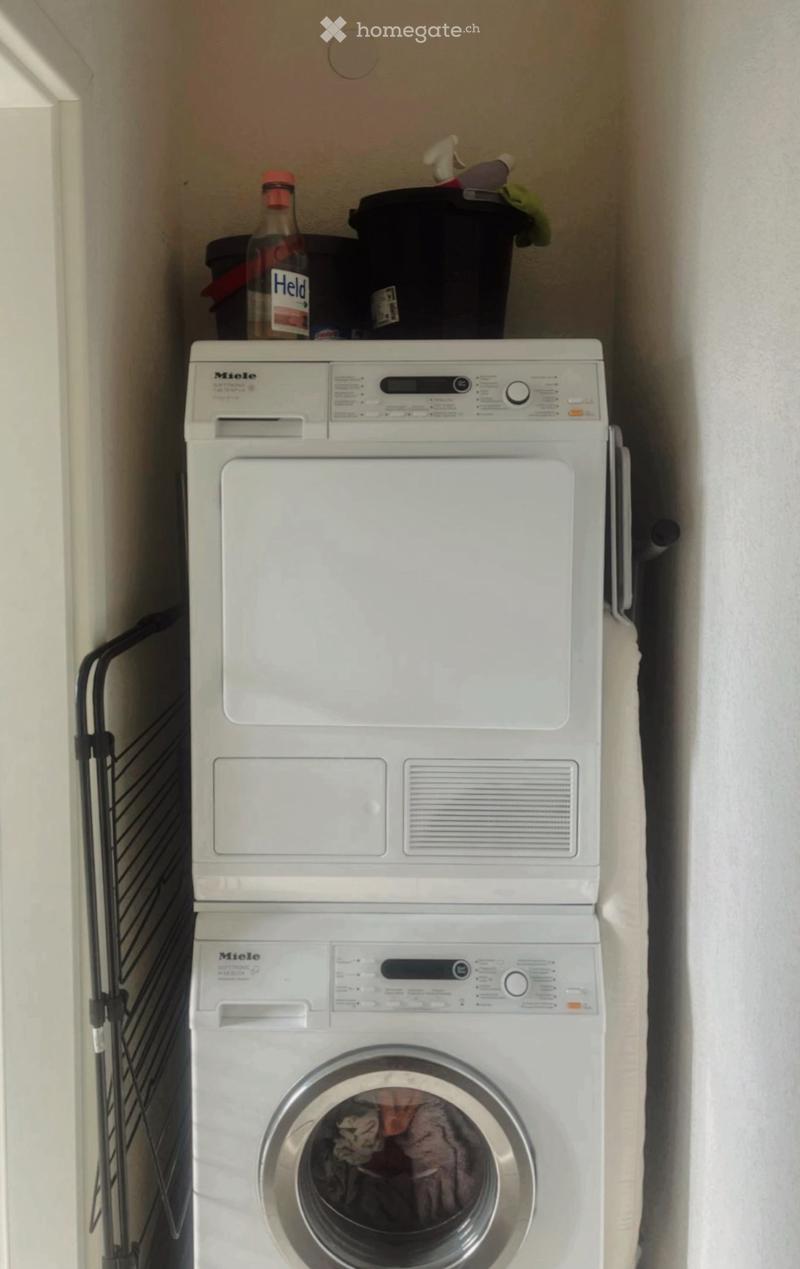 A compact bathroom with a stacked Miele washing machine and dryer unit, a bottle on top, and a small black trash bin.