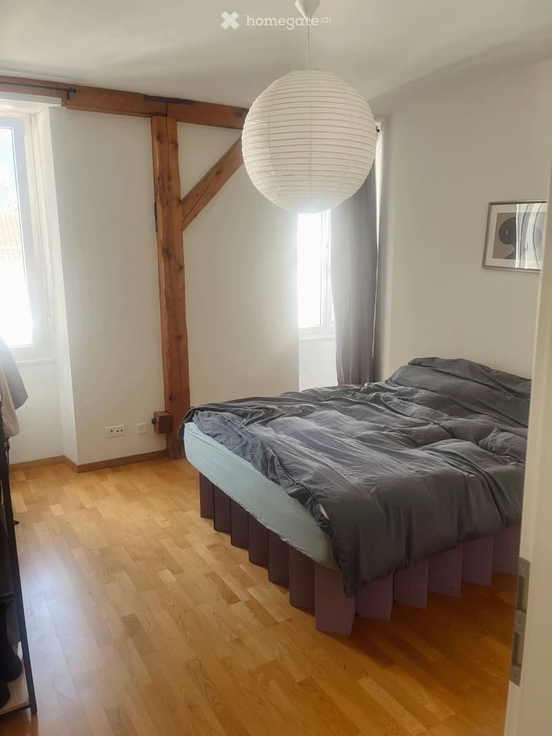 Cozy bedroom with wooden floor, large bed with dark bedding, pendant light, and exposed wooden beams.
