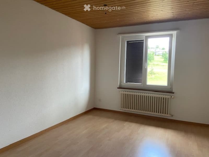 Empty room with wooden flooring, white walls, and a single window with blinds. A radiator is placed below the window.