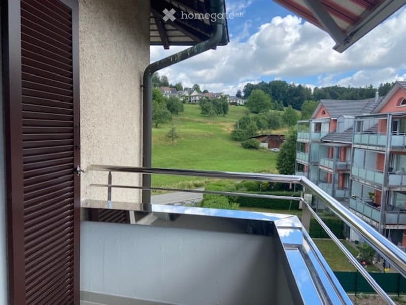 Balcony with metal railing, overlooking a green field and distant houses.