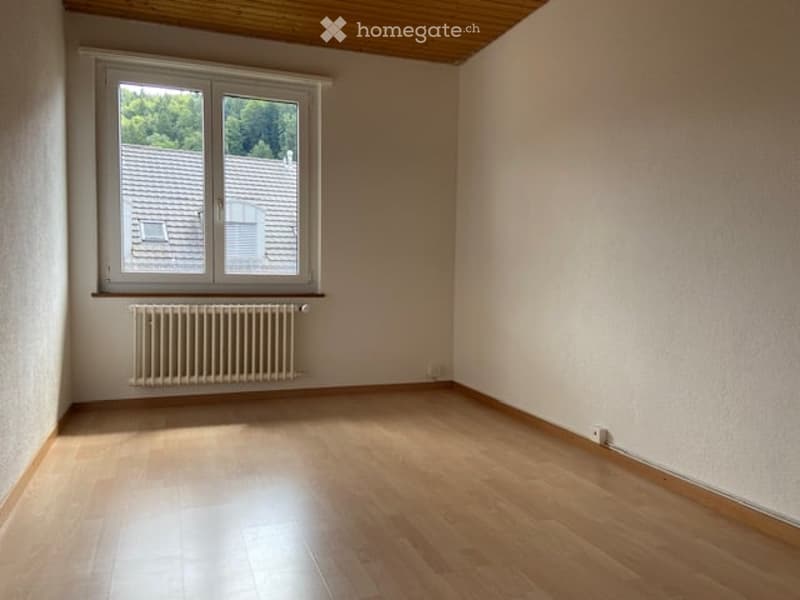 Empty room with wooden flooring, white walls, and a window showing a view of neighboring rooftops. A radiator is mounted below the window.