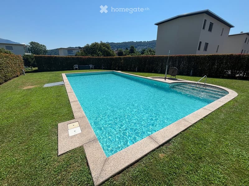 Large rectangular swimming pool, diving platform, hedges, grassy area, white building in the background, mountains in the distance