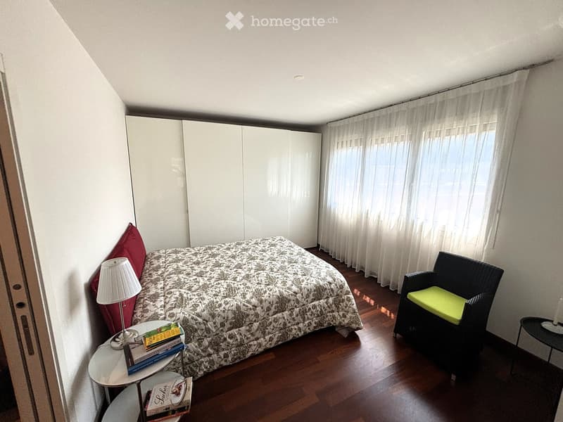 modern decor, wooden floor, large bed, chair, night lamp, table with books, white curtains