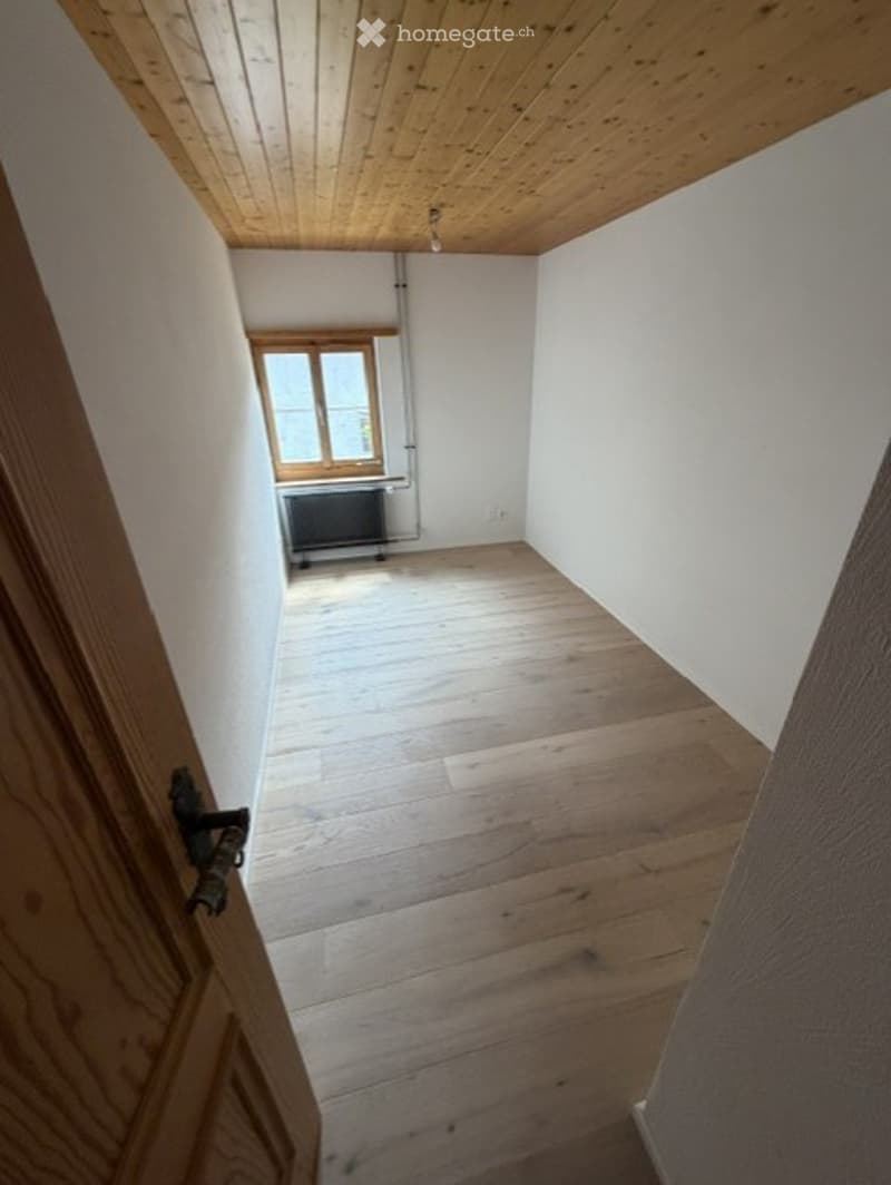 Empty room with wooden flooring and white walls, featuring a wooden door frame and a window with wooden shutters.