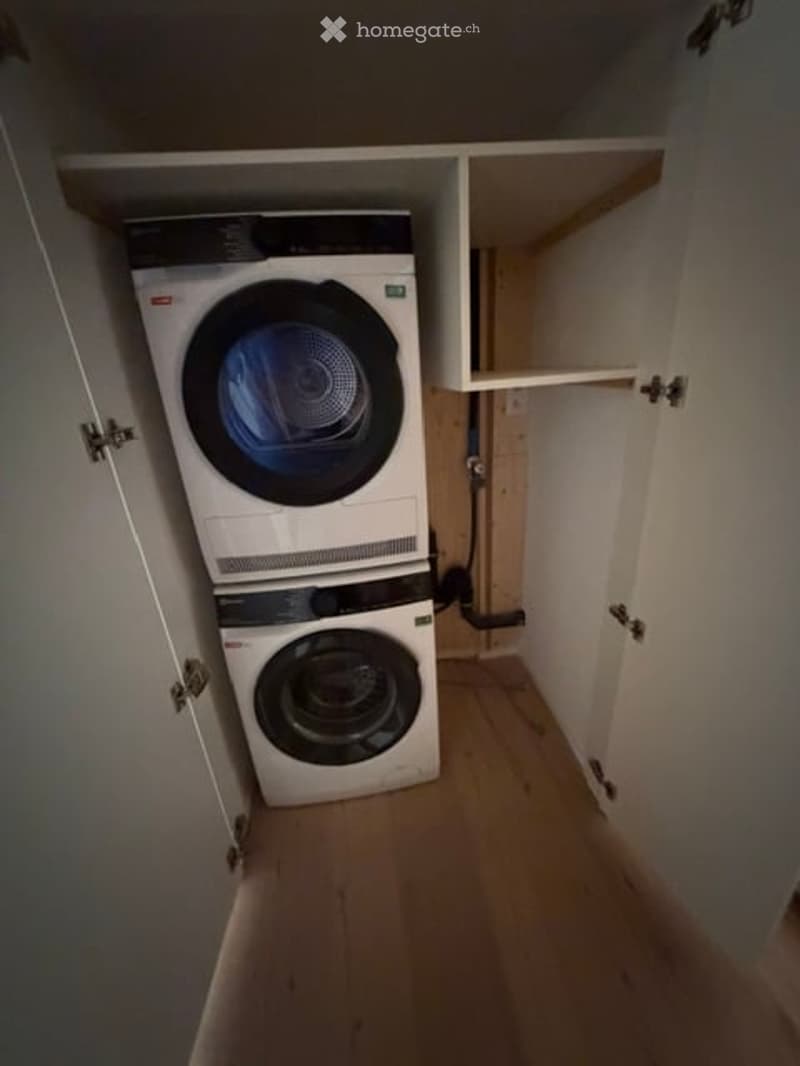 A small, well-organized laundry area with a washing machine and dryer stacked vertically, white cabinets, and wooden flooring.