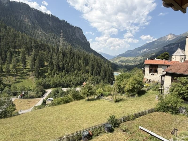 Panoramic view of a lush green valley surrounded by mountains, with a small pond and scattered trees. A rustic building is visible on the right side.