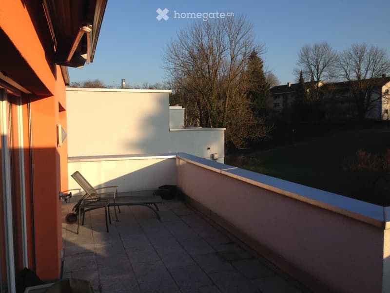 Balcony with tiled floor, white railing, lounge chair, and small table. View of trees and neighboring buildings.