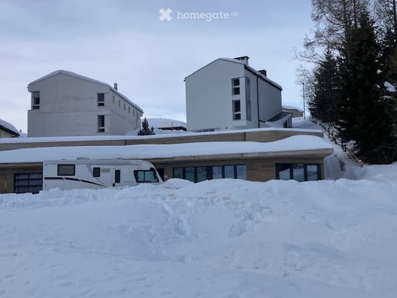A multi-story building with a snow-covered roof and surrounding snow-covered ground. The building has large windows and a visible RV parked in front.