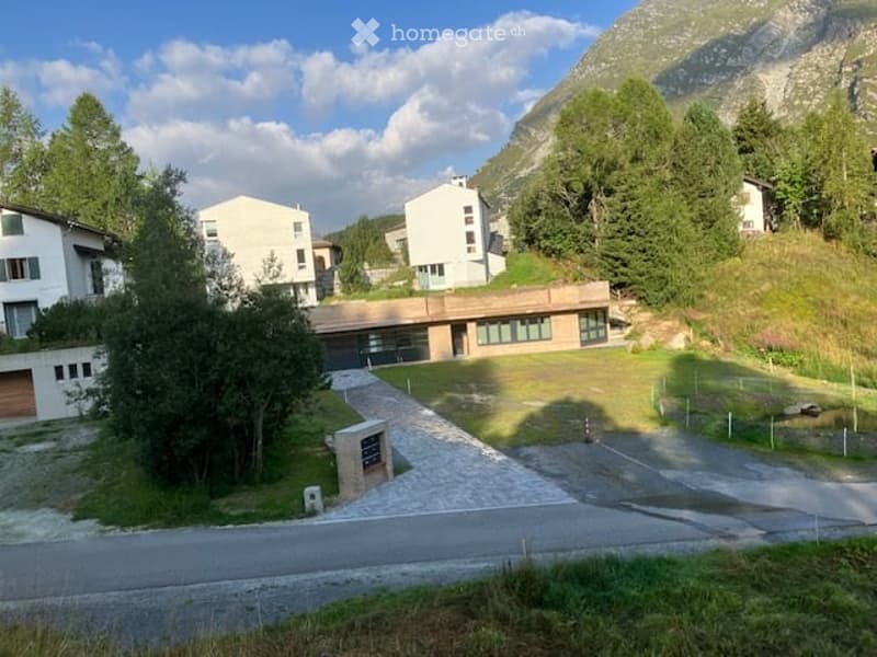 A modern residential complex with multiple buildings, surrounded by trees and mountains, featuring a paved driveway and a small landscaped area.