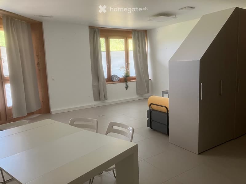 Bright room with white tiled floor, large window with curtains, white table with chairs, grey sofa, and dark cabinet.