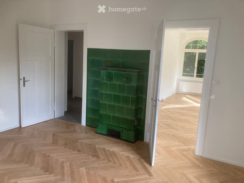 Hallway with wooden herringbone flooring, green tiled fireplace, white walls, and open doors leading to adjacent rooms.
