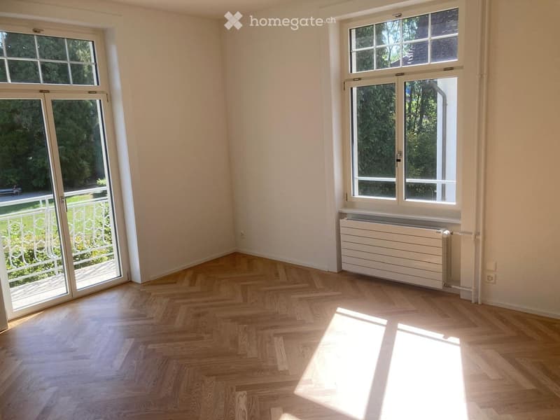 Empty room with wooden herringbone flooring, large windows, and a radiator. The room has white walls and a sliding glass door leading to a balcony.