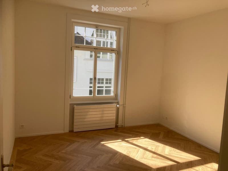 Empty room with wooden flooring, large window with double glass panels, radiator below the window, and light-colored walls.