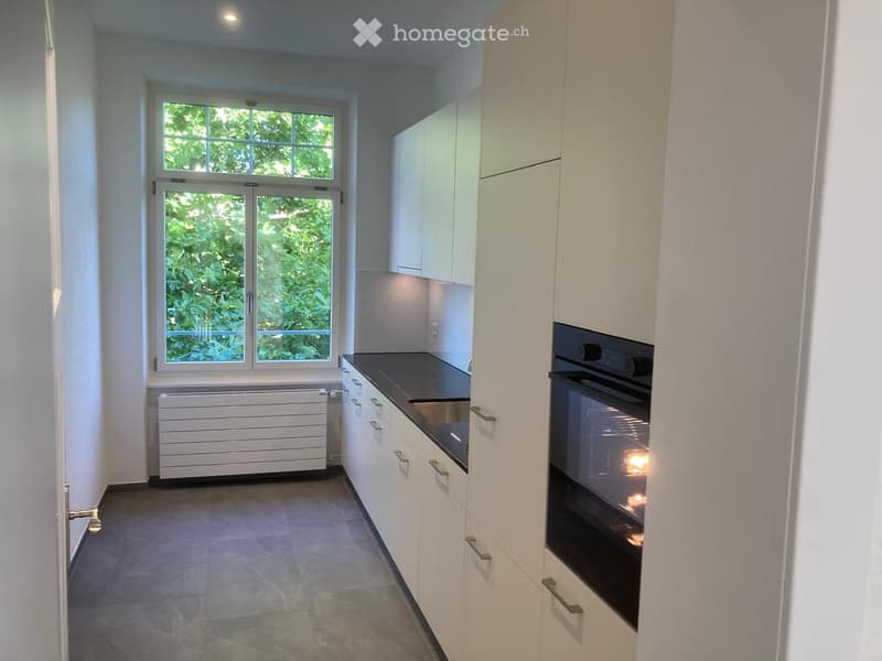 Modern kitchen with white cabinets, dark countertop, built-in oven, and large windows overlooking greenery.
