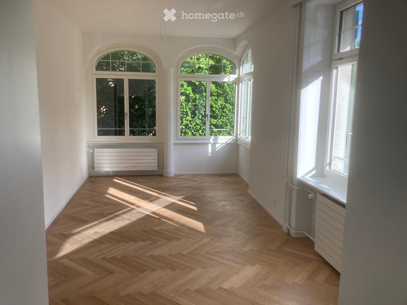 Empty room with light wooden herringbone flooring, white walls, large windows with arched tops, and radiators along the walls.