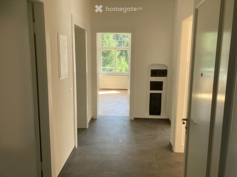 A long, narrow hallway with tiled flooring, white walls, and several doors leading to other rooms. A window at the end provides natural light, and a small built-in storage unit is visible on the right wall.