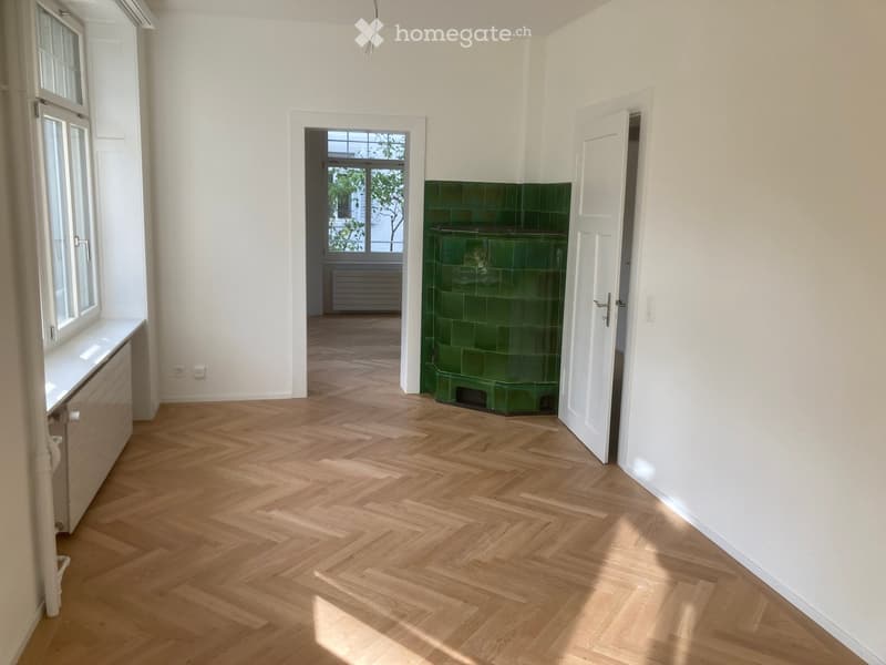 Empty room with herringbone wooden flooring, white walls, and a green tiled fireplace. There are two open doors leading to other rooms, and a window with a bench on the left side.