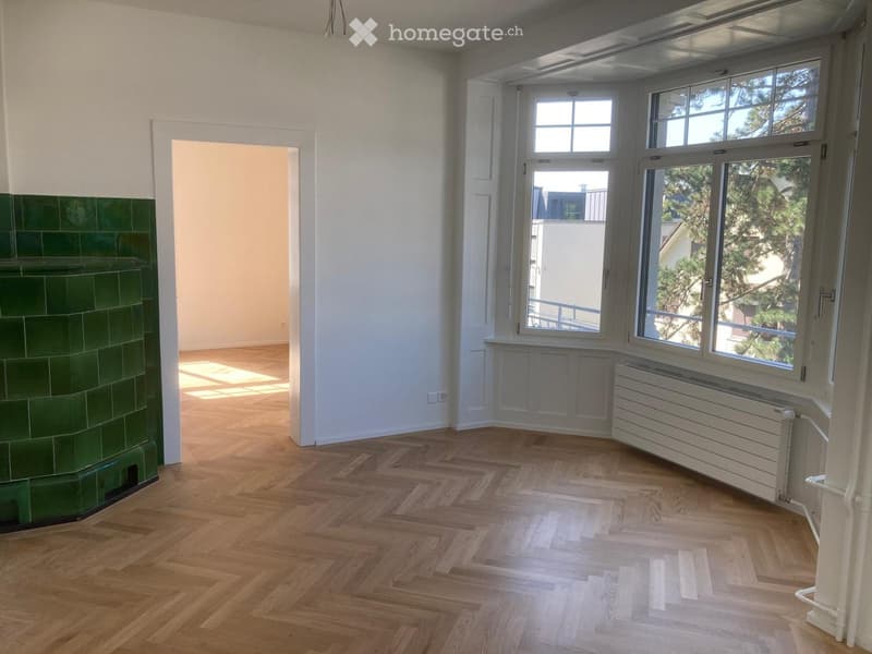 Empty room with wooden flooring, white walls, large windows, and a green-tiled fireplace. No furniture or equipment present.