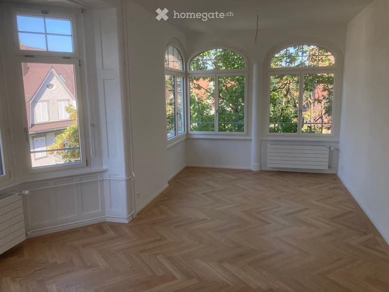 Empty room with wooden herringbone flooring, large windows, white walls, and radiators.