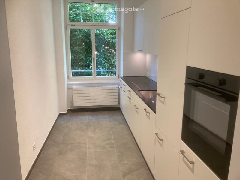 Modern kitchen with white cabinets, dark countertop, and a built-in oven. Features a large window with a view of greenery.