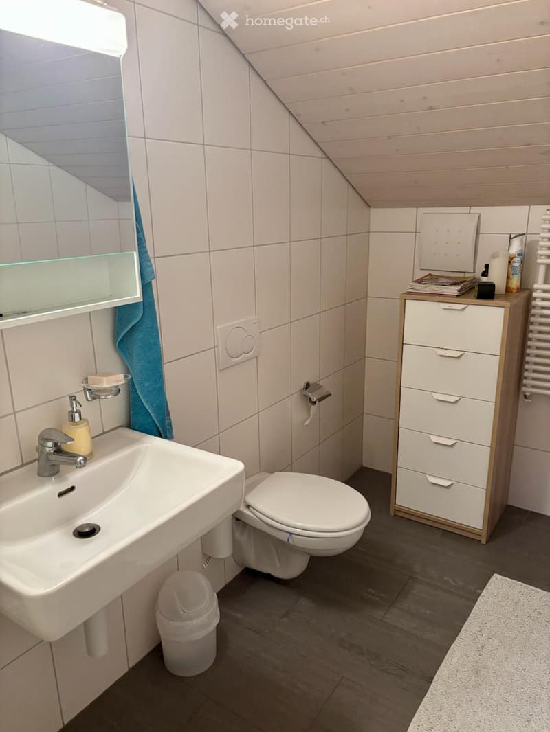 Modern bathroom with white tiles, a sink, toilet, mirror, towel, and a small storage unit with magazines and toiletries.