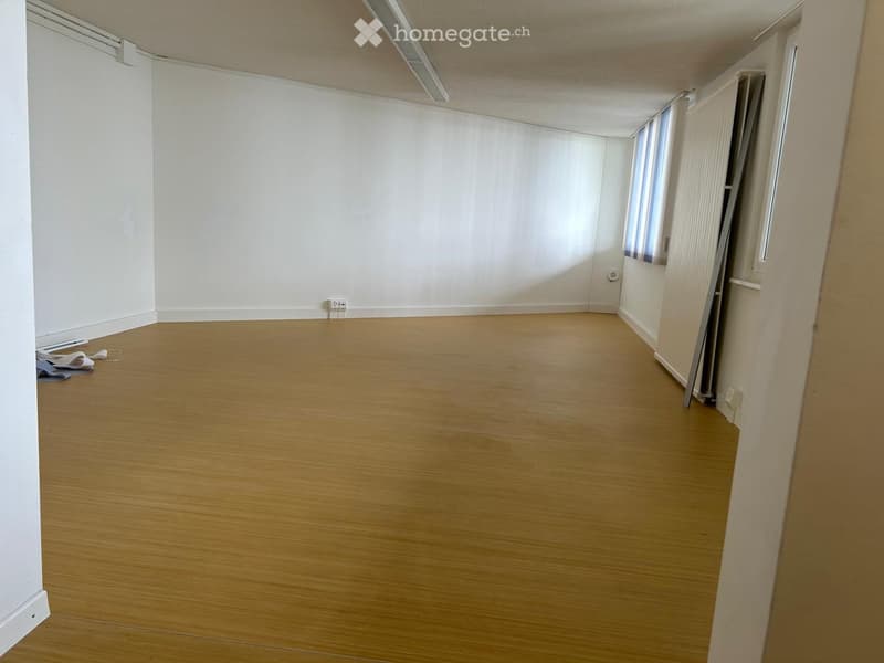 Empty room with wooden flooring and white walls, featuring a window with blinds and a door frame visible.